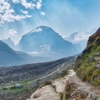 Sun rays break through clouds to shine down on mountains on Plain of Six Glaciers trail and striped cliffs on sunny smoky day