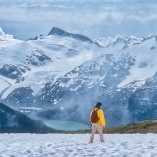 Male hiker treks through snowfield toward alpine lake with snowy mountain ranges in Garibaldi Provincial Park on cloudy day